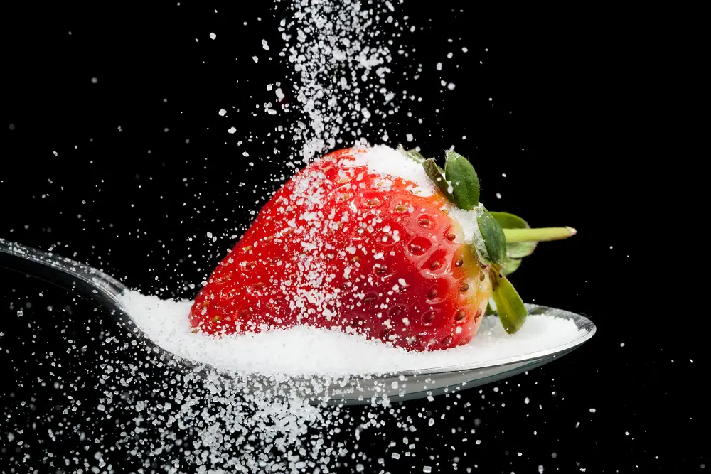 Sugar being poured over strawberry that is sat in sugar on a spoon. This is to represent the amount of sugar in fruit
